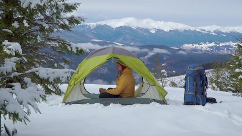 Tourist in a tent in in the mountains in a winter. A man is resting and drinking tea from a yellow mug. Beautiful winter landscape. Travel concept. 4K Arkistovideo