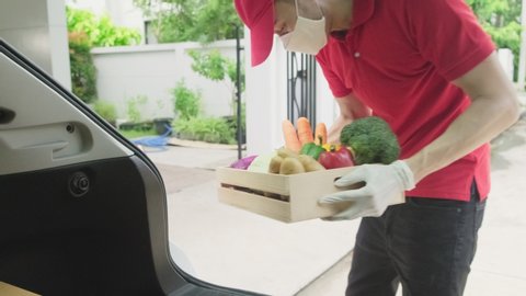 Asian deliver man wearing face mask in red uniform handling box of food, groceries, fruit from car give to costumer in front of the house. Postman and express grocery delivery service during covid19.