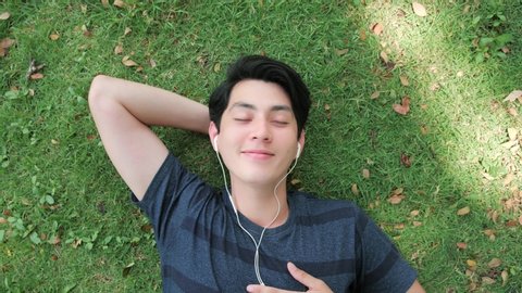 Happy young handsome man relaxing with eyes closed wearing headphones listening to music audio sound on comfortable green grass. Above angle shot.
