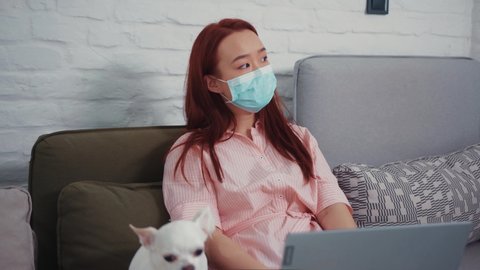 Asian young woman working on freelance project by laptop, wearing protective face mask, looking tired unhealthy. Cute Chihuahua puppy nearby. Staying home.