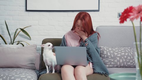 Unwell sick chinese young woman sits with white Chihuahua dog on sofa, working by laptop. Freelance, staying home. Health care. Coronavirus pandemic.