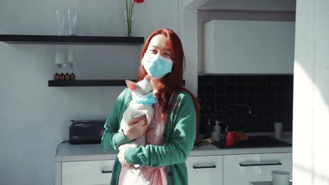 Young calm Asian woman with Chihuahua dog standing in the kitchen, both wearing protective medical masks, staying at home together. Pandemic. Health care. People and pets.