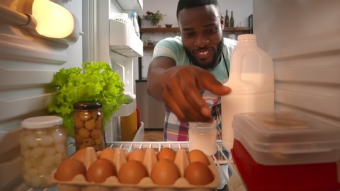 View from refrigerator of young afro-american guy in apron opening fridge and taking ingredients in container cooking dinner. Man taking food from fridge making meal