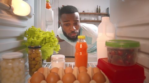 View from fridge of hungry afro-american man opening refrigerator looking for snack and closing in dissatisfied. Upset afro guy mot finding tasty meal to eat in fridge