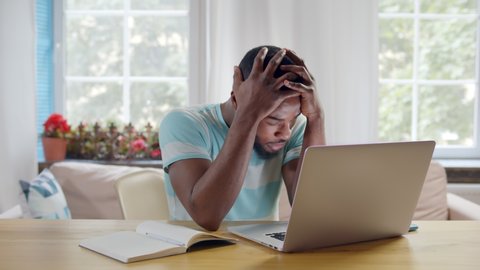 Portrait of afro-american freelancer man thinking about problems and suffering from depression working on laptop at home. Frustrated guy having financial problems