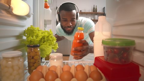 View from fridge of young afro-american guy in headphones taking bottle of carrot juice from refrigerator at home kitchen. Man taking juice from fridge to fresh up
