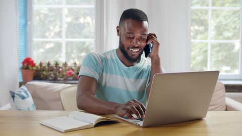 Portrait of afro-american businessman talking on phone with client looking at laptop screen and discussing new startup project working from home office