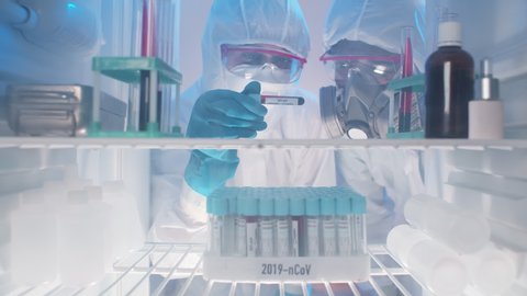 View from fridge of two scientists in protective wear holding blood sample for covid-19 and discussing test results standing near open refrigerator in laboratory