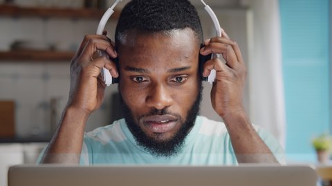 Close up of concentrated young african man in headphones with laptop computer working at home office. Remote job, technology and people concept