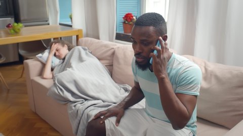 Worried African Husband Calling Doctor While His Wife Feeling Bad Having Headache and Lying On Couch Covered with Blanket At Home. Man consulting with doctor about sick woman