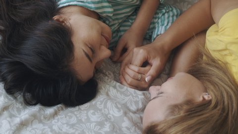 Homosexual mixed race lesbian couple lying on bed, looking each other. Two women sharing love and support holding hands. Gay family. LGBTQI, Pride Event, LGBT Pride Month, friendship concept.