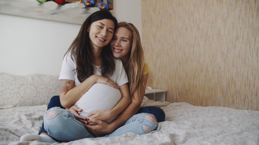 Portrait of happy lgbt lesbian family expecting a baby. Mixed race women in love, petting a big belly. Happiness of motherhood. IVF, LGBTQI, LGBT Pride Month, relationship, childbirth concept. Royalty-Free Stock Footage #1054726622