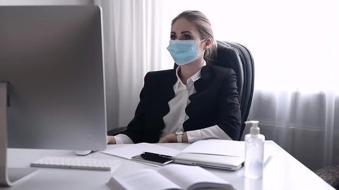 Work during self-isolation and quarantine. Young woman employee in a medical facial mask works in the office at the computer. Coronavirus Pandemic Covid 19.