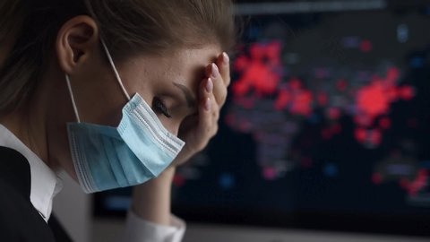 Side view close-up portrait of a young woman in disposable medical facial mask holding her head and looking at the epidemic map of the coronavirus Covid 19 pandemic on a computer