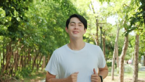 Happy handsome Asian man running in park on public green background, Fit man jogging at outdoor workout on background of trees. Healthy lifestyle concept. Slow motion
