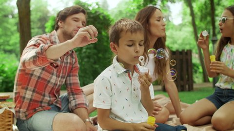 Positive girl and boy playing with soap bubbles in park. Cheerful family having picnic in forest. Parents and kids having fun together outside. Father talking care about son outside