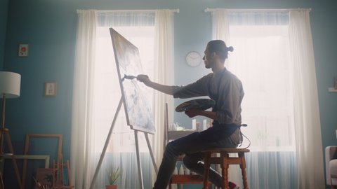 Young artist is staying home, inspired to paint in the morning, sitting on a stool and creating a new artwork, enjoying his creative work that makes him happy, Zoom in, Slow motion.