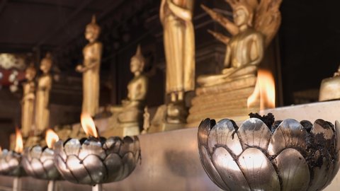 Close-up shot of candles with moving flame in lotus-shapes candle holders with row of golden Buddha statues on the background in a Buddhist temple.