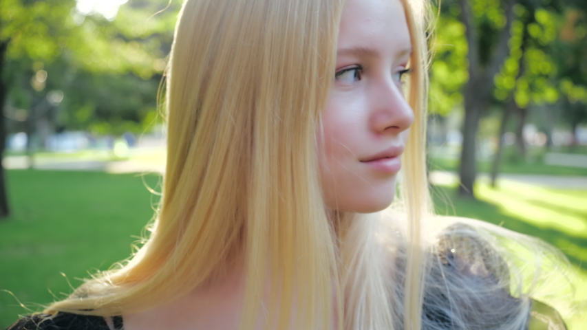 Attractive European blonde teen girl with natural make-up walks in nature on a sunny day. Beautiful fashionable teenage model soft smiles, posing outdoors straightened long hair. People style concept Royalty-Free Stock Footage #1054736660