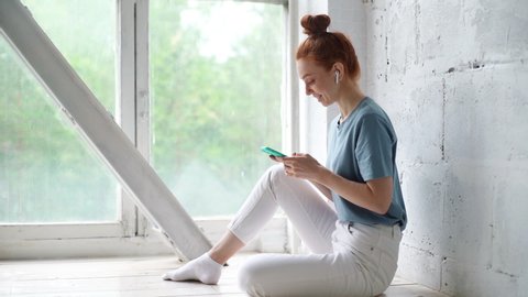 Happy redhead young woman is using cell phone sitting by the big window. Smiling relaxed business lady typing message on cellphone. Cheerful happy girl listens to music with headphones, slow motion.