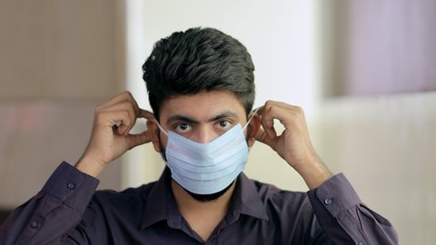 Closeup shot of a young man using a medical mask during the Covid-19 pandemic. Handsome Indian guy wears a surgical mask and shows a thumbs-up sign to protect himself from coronavirus