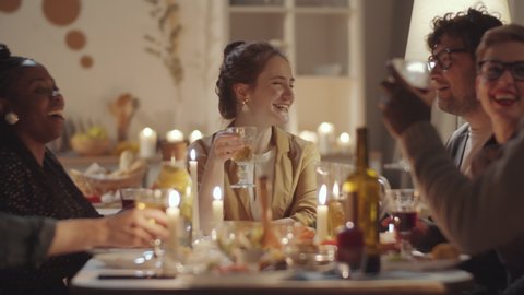Group of multiethnic friends sitting together at dinner table with candles and food on it, clinking glasses together and drinking wine after cheerful woman giving a toast at home party