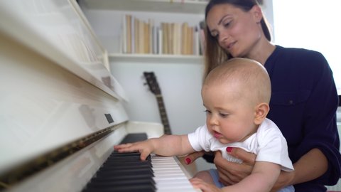 A cute baby boy playing a white piano with his mommy teaching him and holding him in her lap 4K