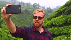 redhaired ginger bearded man in sunglasses talking by smartphone video chat and say hello from india munnar kerala tea plantations