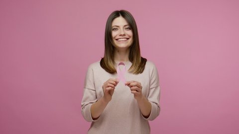Kind brunette woman holding pink ribbon, symbol of breast cancer awareness, recommendation and warning of cancer prevention, female health diagnostics. indoor studio shot isolated on pink background