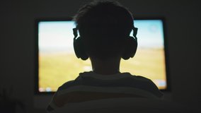 Boy plays game strategy online on computer with headphones in evening in room. Light of monitor, silhouette of child play emotionally at computer. Hobbies of adolescents. Online games on the Internet