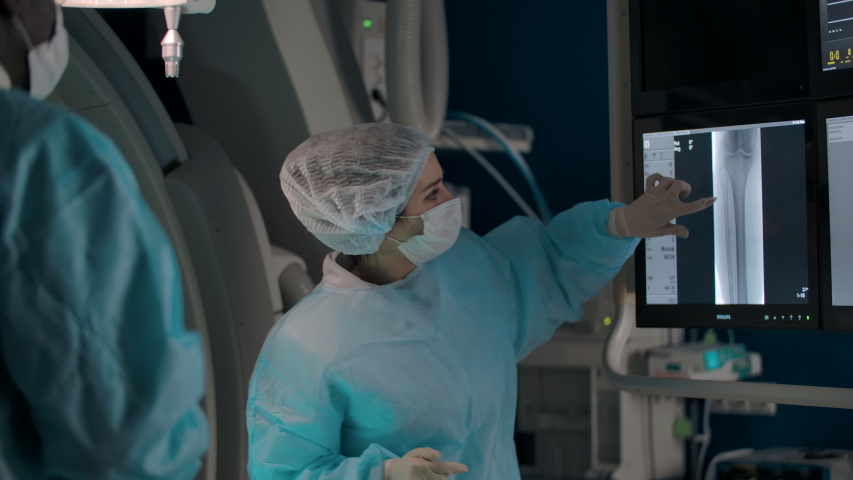 A female surgeon examines an X-ray of her leg with colleagues in the operating room. Her colleagues black and European surgeons are discussing an operation plan | Shutterstock HD Video #1054743932