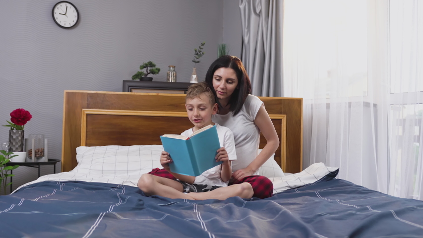 Splendid satisfied 30-aged woman playing with her handsome 8-aged son while he reading interesting book in contemporary sleeping room ,front view