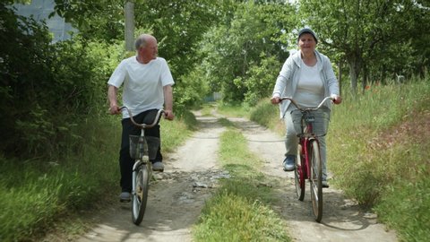 family walk, elderly husband and his beloved old wife enjoy bike ride together on rural road amid green trees on sunny summer day
