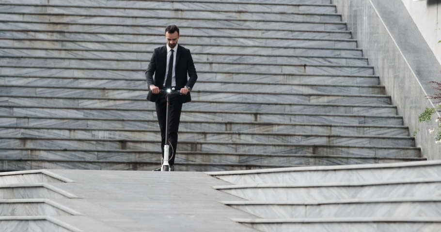 A man of european appearance in a business suit rides an electric scooter, driving away from the stairs down the street.