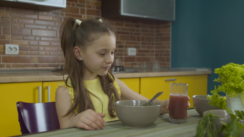 Cute little girl with two ponytails sitting at the table in the kitchen, drinkin juice and eating vegetarian salad. The child is having fun and smiling. The concept of healthy eating
