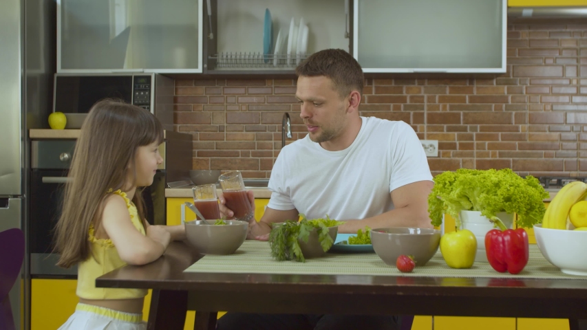 Young happy family dad and little daughter enjoy dinner together in the kitchen. Father talking and having fun with daughter, drinking juice. Girl eating salad