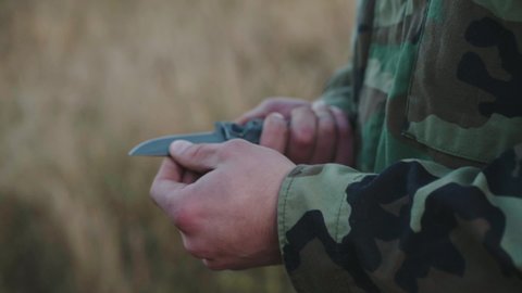 Hands of a fighter in camouflage use a pocket knife outdoors. Slowly