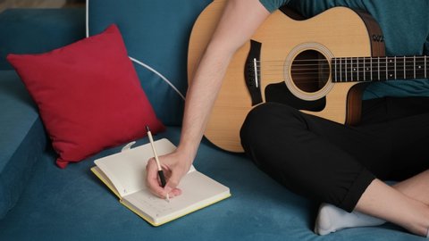 Close up of a guitarist hands that is sitting on blue sofa with a guitar in hand and write new song idea in a notebook