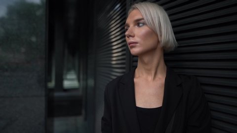 Close-up portrait of a young business girl with short white hair, she walks against a city background and against a background of black shop Windows. She's wearing a black jacket. Poses.