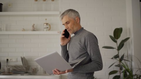 Senior man talking on cell phone pacing back and forth at home with laptop in his hands. Businessman finishing call, sitting down at desk, working on pc and thinking. Online and remote work concept