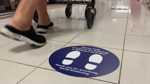 A low angle view of a social distance reminder sticker on the floor of a department store as an elderly woman pushing a shopping cart walks past.	
