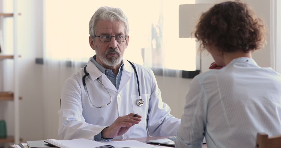 Serious focused middle aged hoary physician in medical coat sitting at table, consulting female patient about illness or surgery. Rear view young woman listening to old doctor at checkup meeting. | Shutterstock HD Video #1054771361