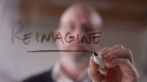 Rack focus of a modern businessman writing "Reimagine" on clear glass