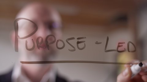 "Purpose-Led" written on clear plexiglass with a dry erase marker by a modern businessman