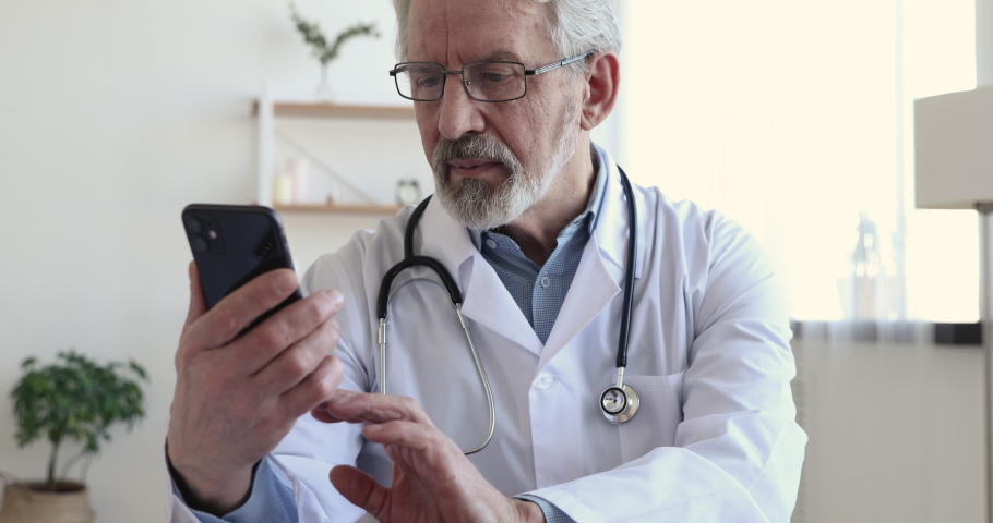 Happy senior old male doctor in medic coat and eyeglasses using smartphone, sitting at workplace. Focused middle aged physician consulting clients online via mobile healthcare application in office. | Shutterstock HD Video #1054774286