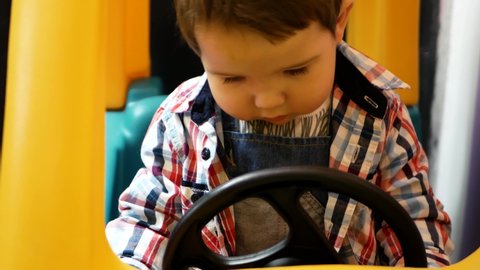 A child plays at the wheel of a toy car. Little cute boy sits in a beautiful, bright children's car at the wheel in the children's play center. Turns the steering wheel, smiles and laughs 0-1 year