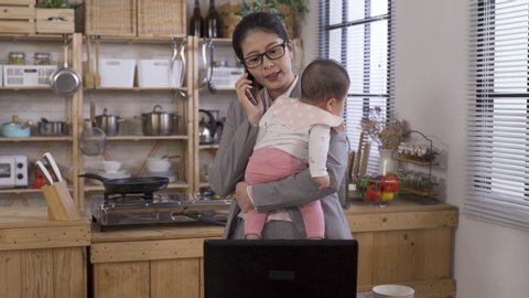 busy taiwanese mom standing with baby is dealing with work on phone. asian career woman holding an infant in front of laptop is looking at data and explaining via cell phone. work from home concept