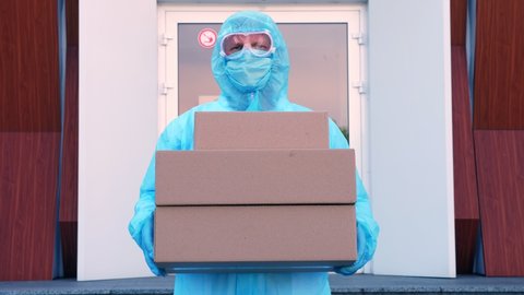 Courier, in protective uniform, mask, goggles, holds in hands some cardboard boxes, on background of entrance door to hospital. Cargo delivery service during coronavirus outbreak.