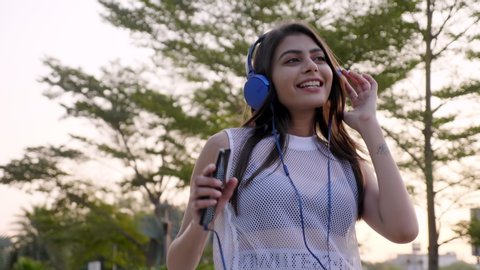 A stylish, fashionable, trendy young woman or female wearing headphones and listing to online music using a smartphone. A beautiful Indian girl enjoying and dancing to an online upbeat song outdoors