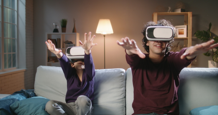 Funny south asian siblings or father and son with curly hair are trying virtual reality headsets, having fun playing video games - modern technologies, family time concept 4k footage